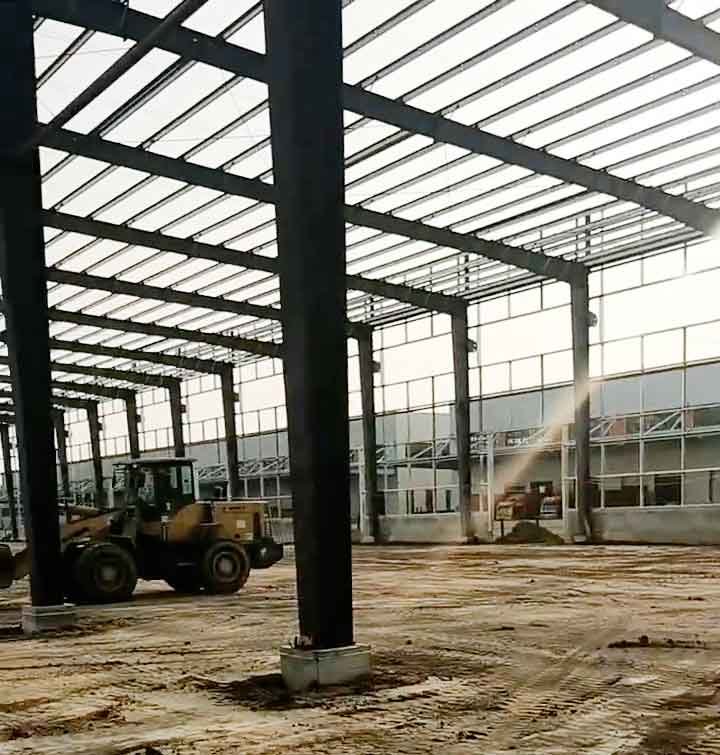 Un cargador frontal amarillo realizando trabajos de movimiento de tierras en el interior iluminado de la estructura de grandes naves industriales metálicas.
