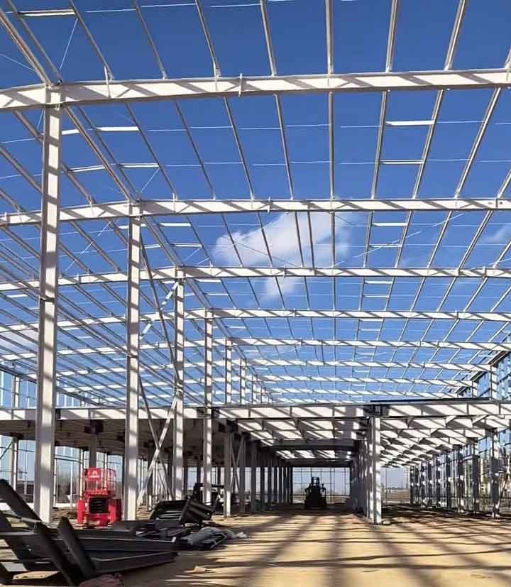 Esqueleto estructural interior de naves industriales proyectando intensas sombras geométricas sobre el suelo de arena, con un área de dos pisos a la derecha y un montacargas al fondo.