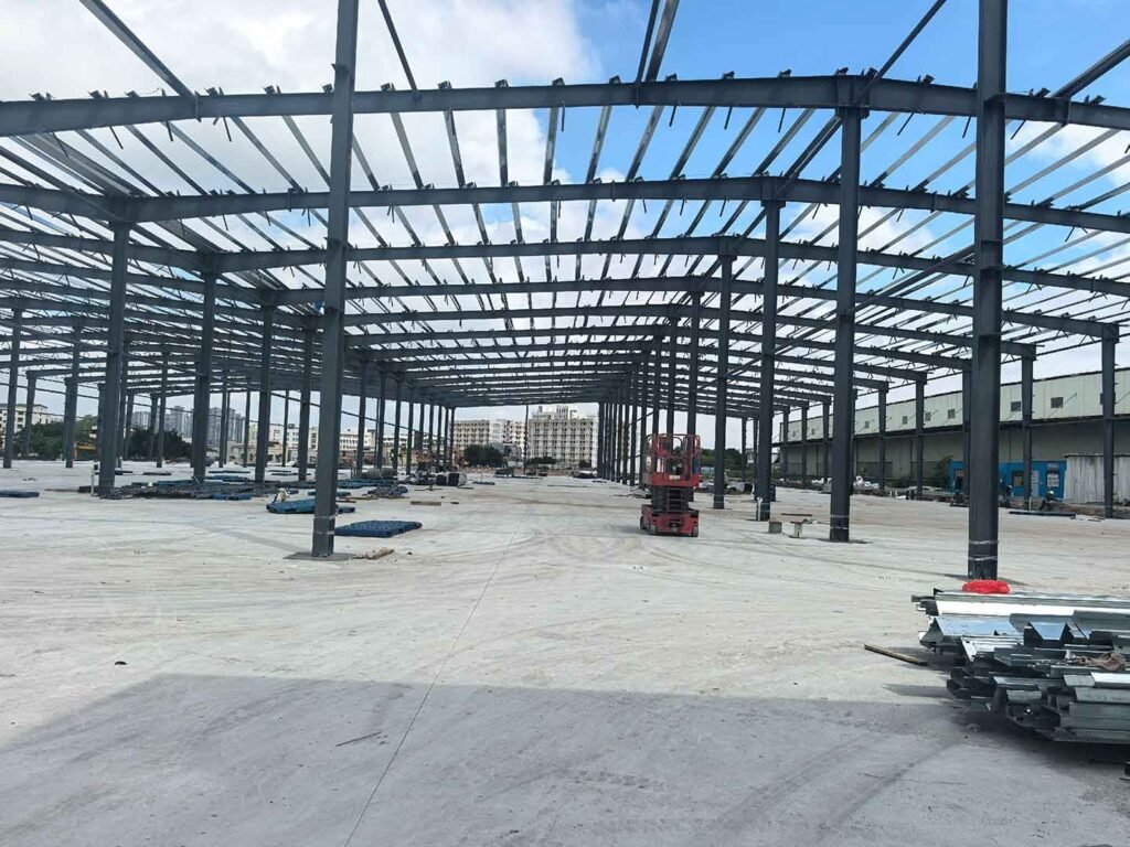 Amplio interior de nave industrial mostrando densas estructuras metálicas en el techo, una plataforma elevadora roja y perfiles galvanizados apilados en primer plano