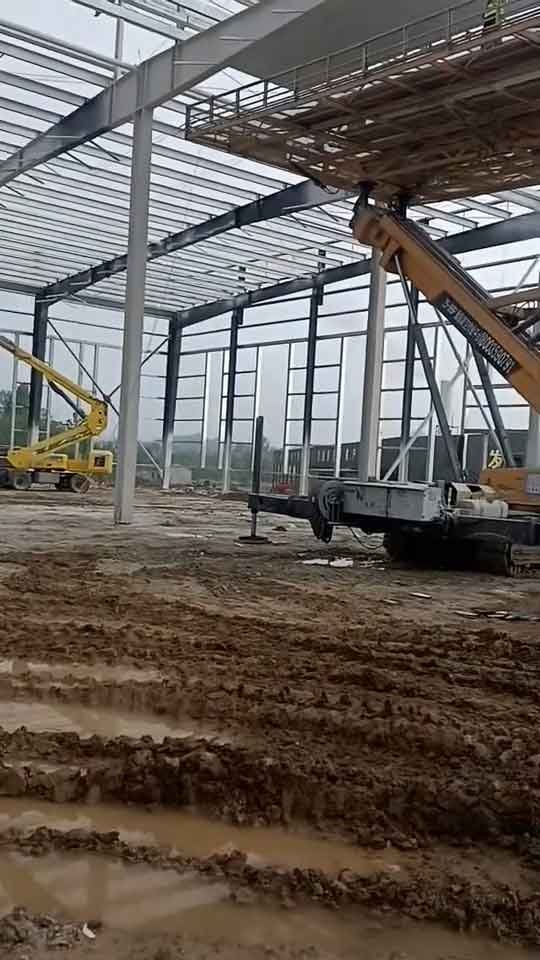 Sitio de obra mostrando una construcción industrial económica con estructura metálica blanca, un rodillo compactador amarillo trabajando y barriles negros en el suelo.