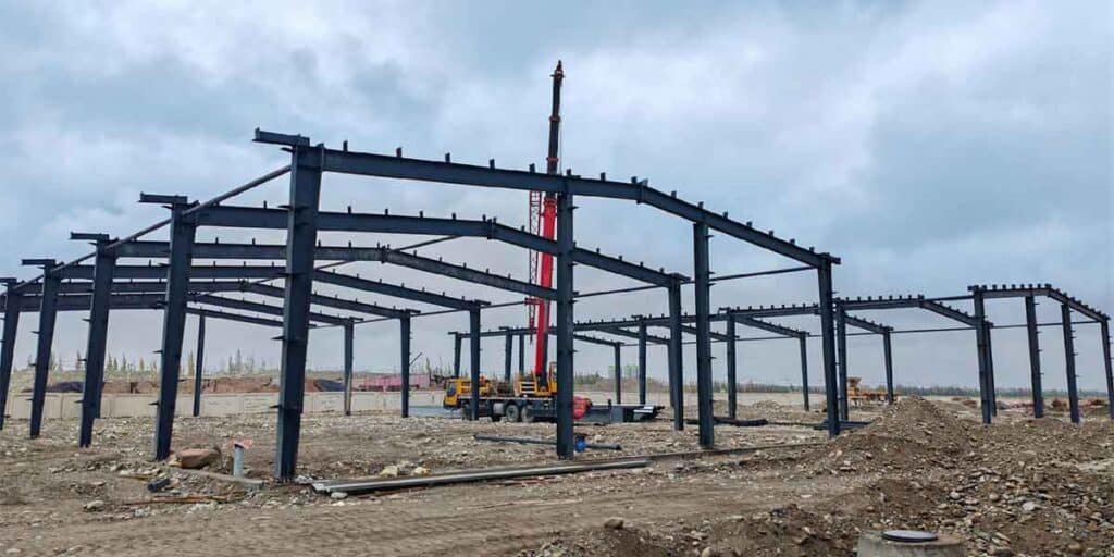 A red mobile crane hoisting heavy black structural roof beams on an overcast day, utilizing precise components from a professional prefabricated steel warehouse supplier.