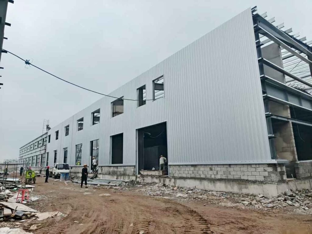 Larga fachada exterior de una gran bodega industrial casi completamente revestida con paneles de acero corrugado gris claro, con obreros en la base.