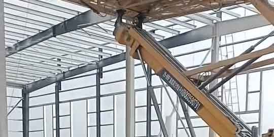 Brazo mecánico de una plataforma elevadora amarilla sosteniendo componentes para soluciones en acero en el techo de una estructura metálica blanca.