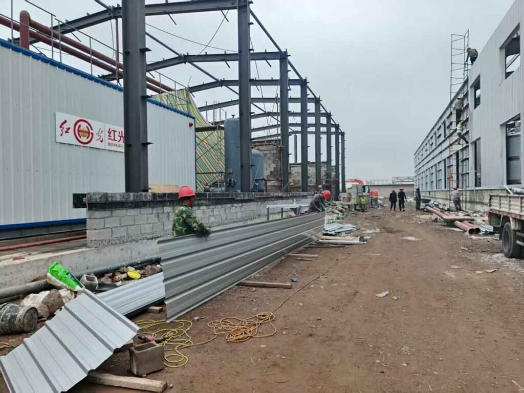 Construction workers moving a very long corrugated metal sheet on the ground level of a steel warehouse site with exposed black steel columns.