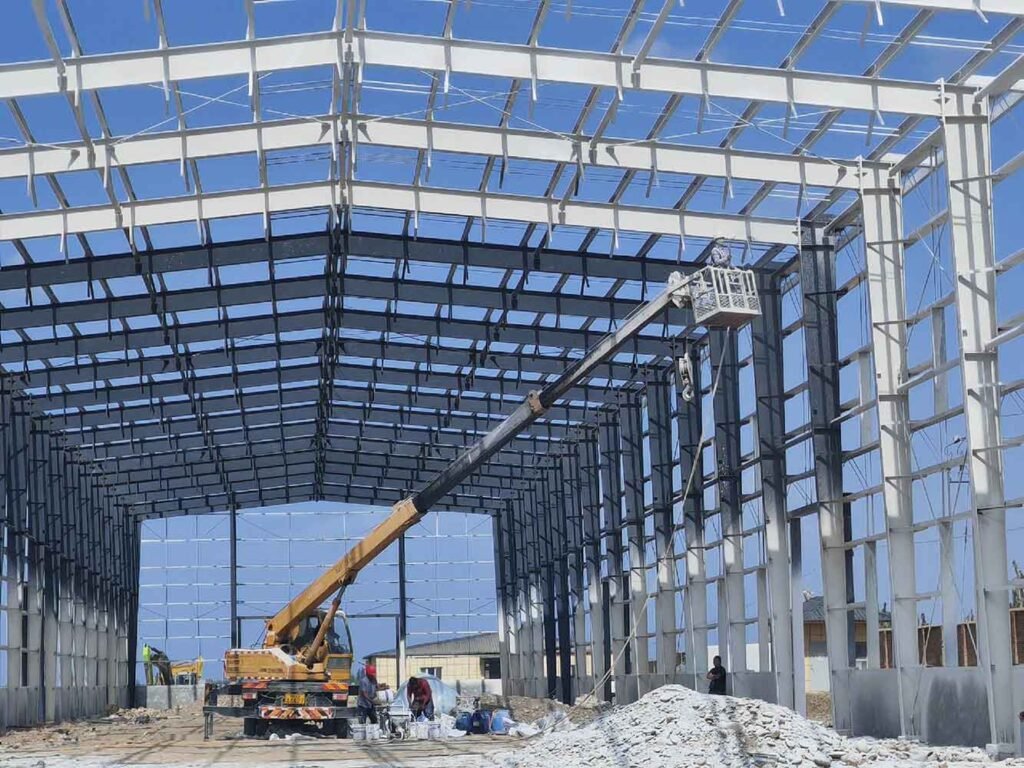 Construction workers in a yellow crane basket adjusting white painted roof purlins above ground material staging for a bright steel warehouse Colombia