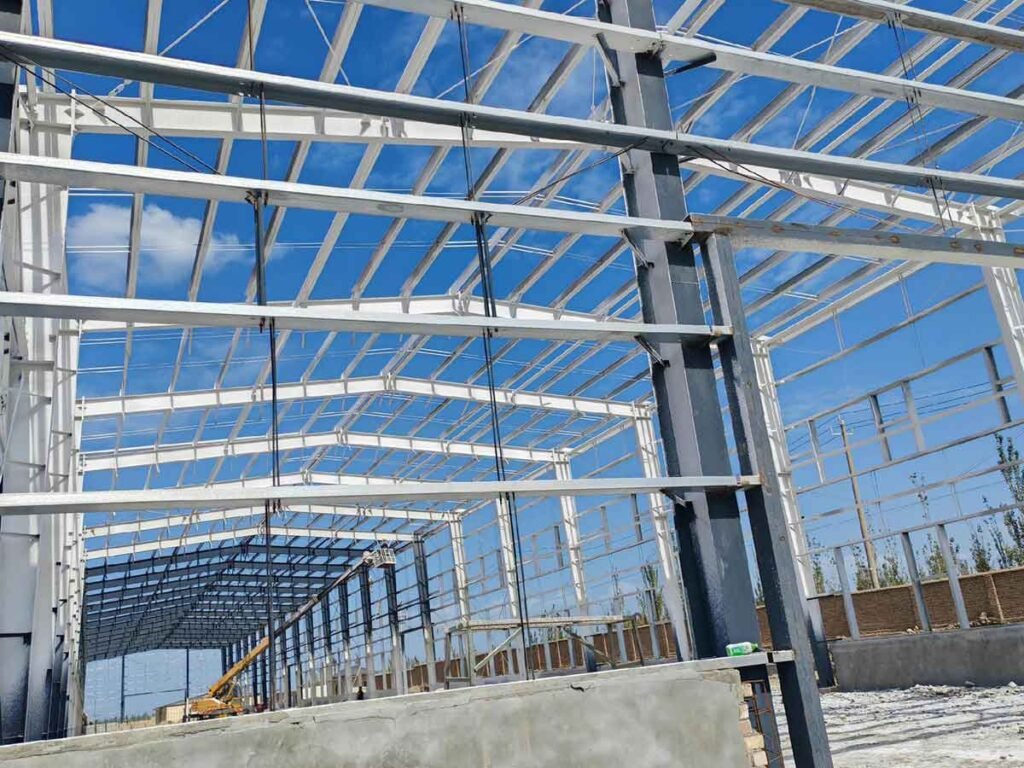 High-altitude construction workers balancing on black structural beams assisted by a ground worker with a rope, assembling a heavy-duty steel structure warehouse and industrial warehouse Colombia.