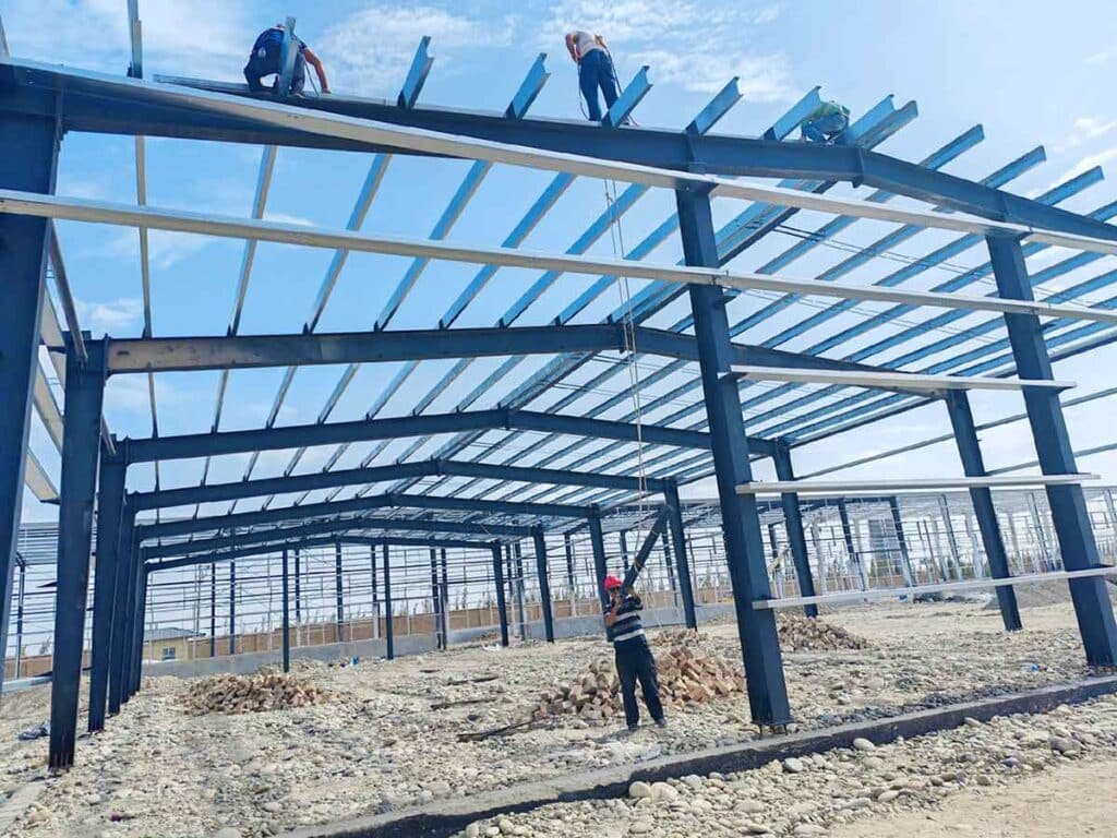 Skilled construction workers balancing on high black roof beams to install white purlins, assisted by a ground worker with a rope over a cobblestone floor for a sturdy steel structure warehouse.
