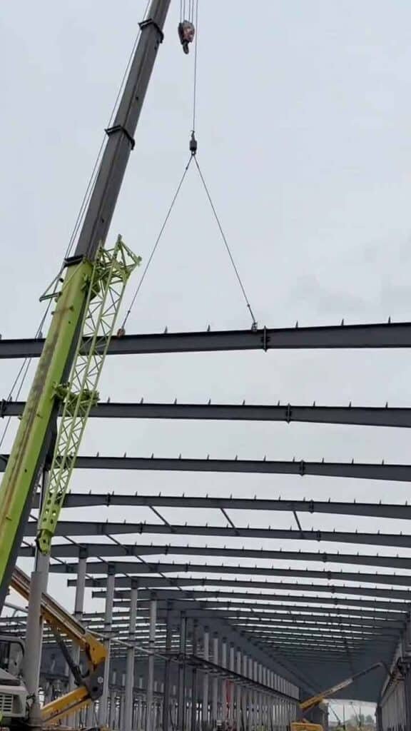 Brazo telescópico de una grúa verde bajando una viga de acero horizontal mediante cables con ganchos hacia la estructura superior del techo de un almacén metálico en construcción.
