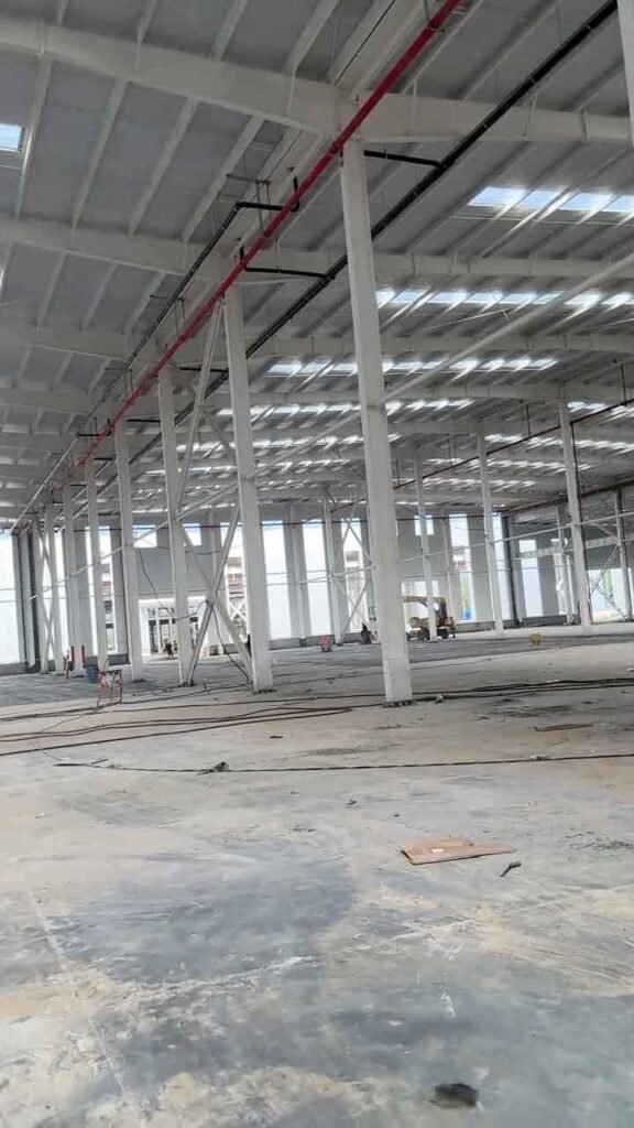 Interior de un almacén prefabricado mostrando tensores diagonales en X en el lateral izquierdo para refuerzo estructural, vigas continuas y maquinaria de construcción amarilla al fondo.