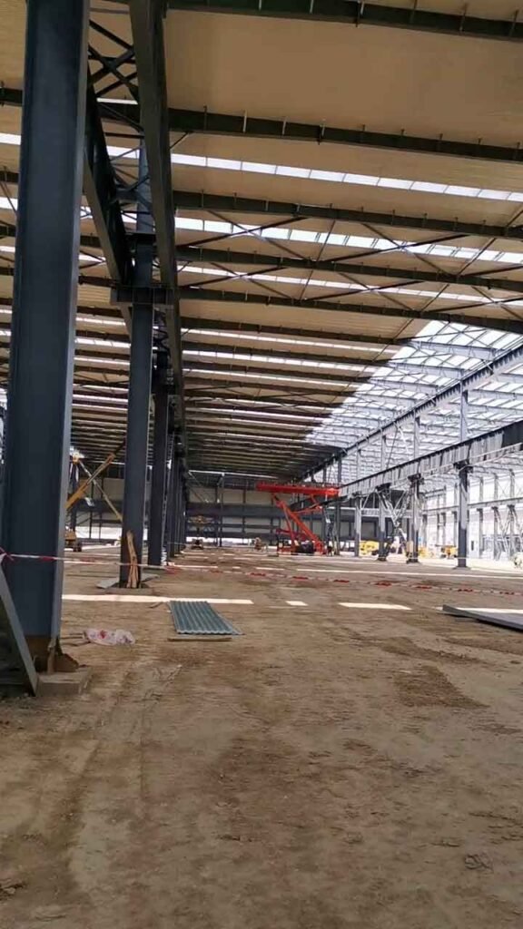 Interior profundo de una nave industrial moderna en construcción, mostrando columnas de acero oscuro, techo con tragaluces y cinta de precaución en el suelo de tierra.