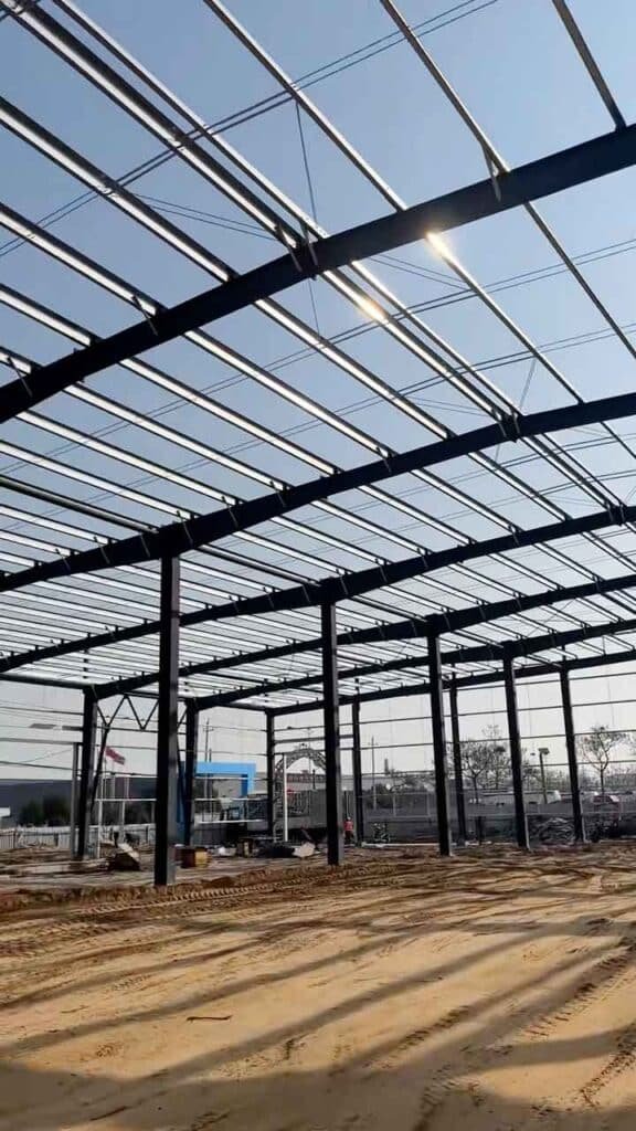 El sol brilla a través del techo abierto de estructuras metálicas modernas proyectando sombras lineales sobre el suelo de arena nivelado.