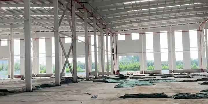 Muro perimetral de una bodega industrial prefabricada con columnas de acero y refuerzos en cruz, grandes aberturas rectangulares que muestran árboles verdes en el exterior, y plásticos en el suelo.