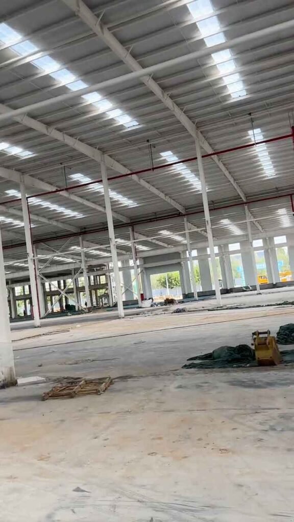 Vista interior de una bodega metálica en construcción, destacando columnas de acero blanco, techo con tragaluces de luz natural y sistema de tuberías rojas contra incendios sobre un piso de concreto