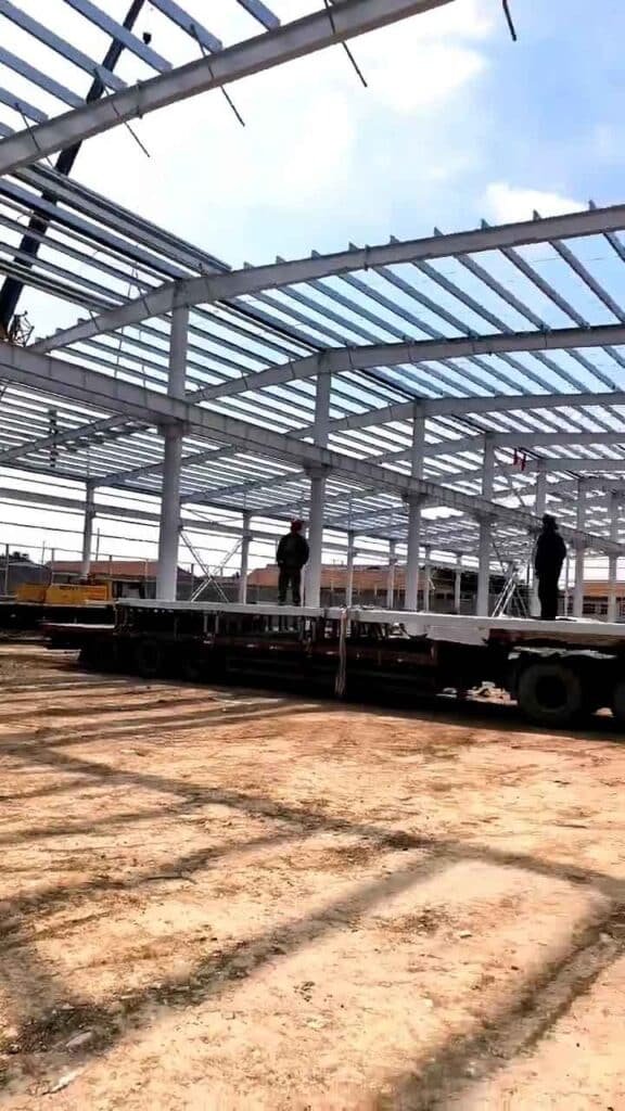 Camión plataforma facilitando la construcción industrial eficiente mediante la descarga estratégica de perfiles de acero pre-pintados.