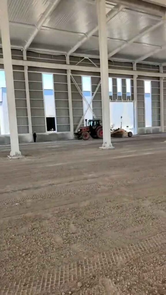Maquinaria trabajando en la base de una estructura metálica industrial con vigas de soporte blancas y techo galvanizado.