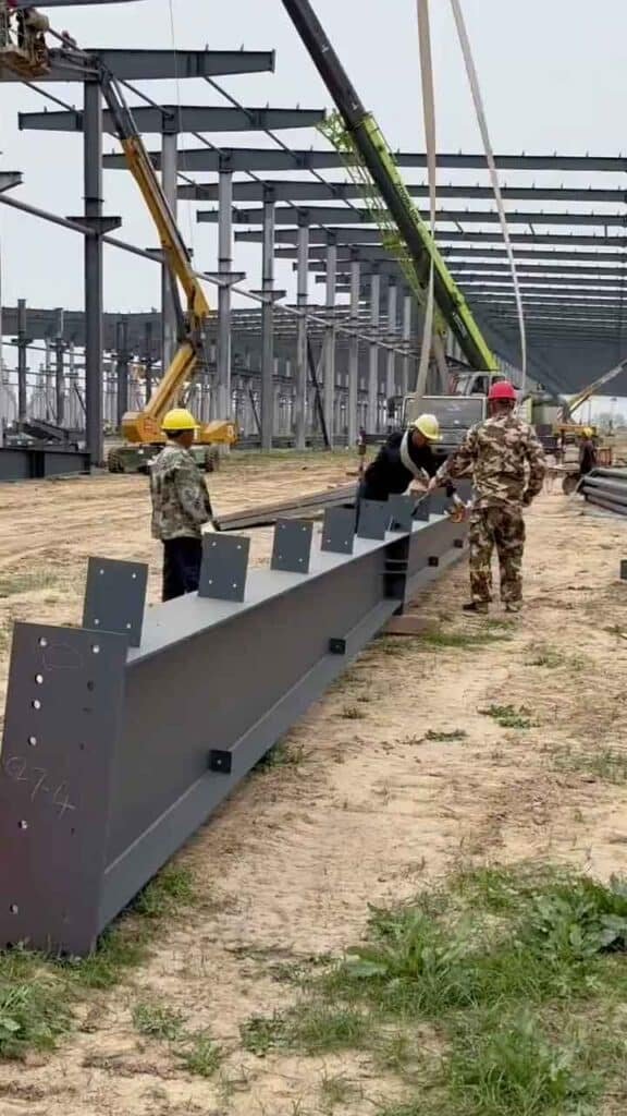 Trabajadores con cascos de seguridad y ropa de camuflaje coordinando maniobras con una grúa verde en la fase inicial de construcción de un gran galpón industrial sobre terreno de tierra.