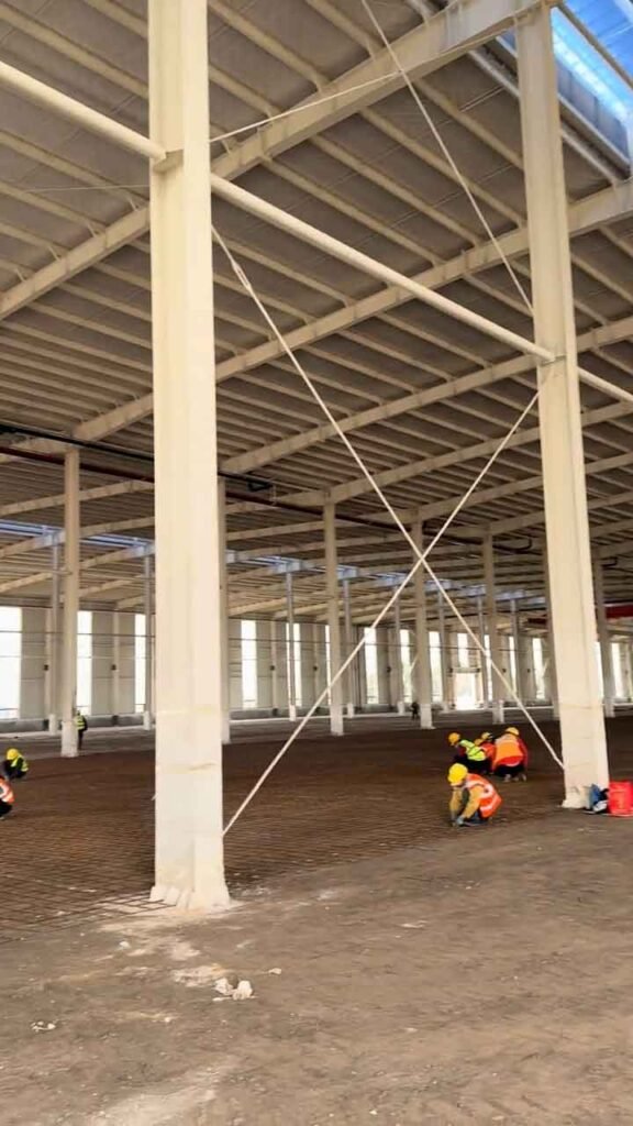 Trabajadores instalando refuerzos en el suelo de un galpón industrial metálico con tensores de acero en los pilares