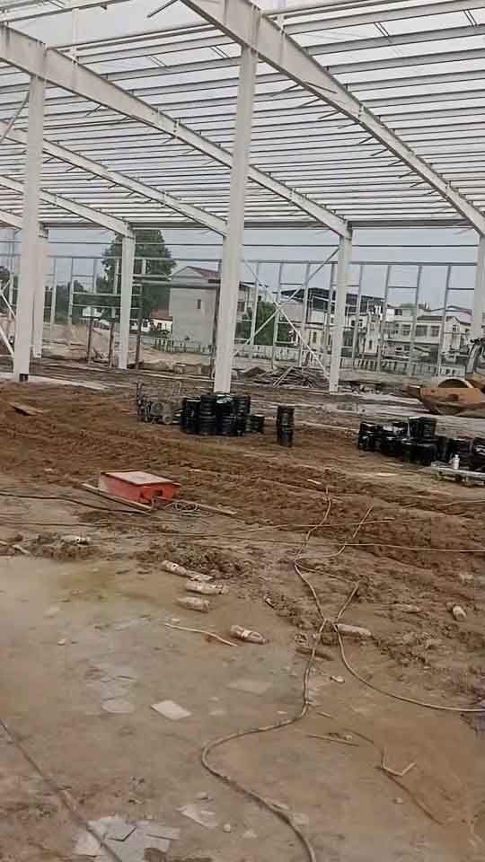 Estructura de un galpón industrial prefabricado en etapa de montaje con materiales de cimentación y bidones de sellante en primer plano.