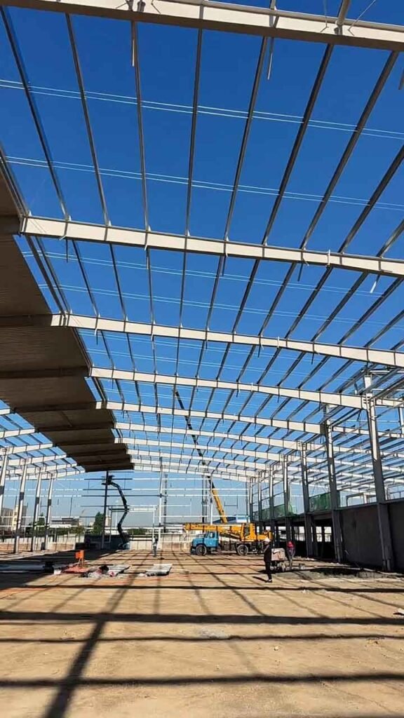 Obra en construcción de un galpón metálico de grandes dimensiones con grúas y camiones operando.