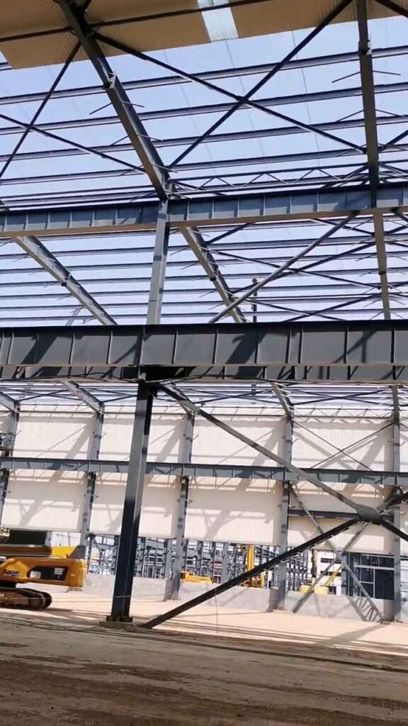 Esqueleto metálico oscuro de almacenes prefabricados mostrando el avance en los cerramientos de pared blancos al fondo y maquinaria de obra a la izquierda.