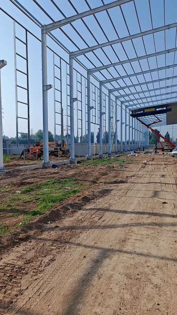 Estructura de una nave industrial metálica en fase de montaje con pilares de acero blanco y vigas de techo sobre terreno compactado.