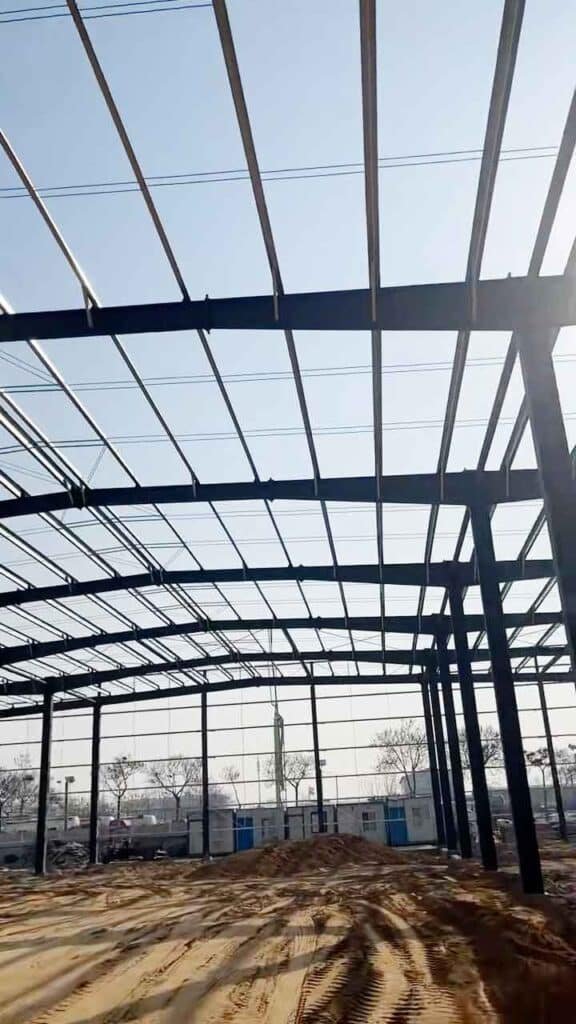 Esqueleto de acero oscuro de una nave industrial modular en construcción con cabinas de contenedores azules al fondo.