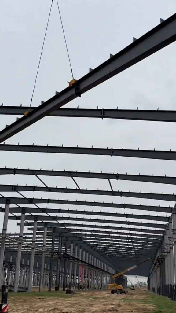 Vista profunda del eje central de una nave prefabricada, mostrando maquinaria pesada al fondo y una larga viga de techo suspendida en el aire por cables durante su instalación.