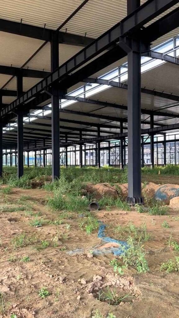 Columna de acero negro en primer plano sosteniendo el techo con tragaluces de una nave industrial modular sobre tierra con un plástico azul.