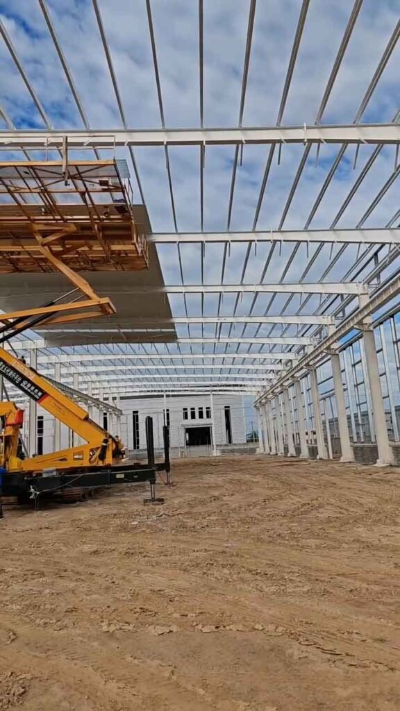 Montaje de cerchas de techo en un galpón industrial de acero con maquinaria de elevación amarilla en terreno preparado