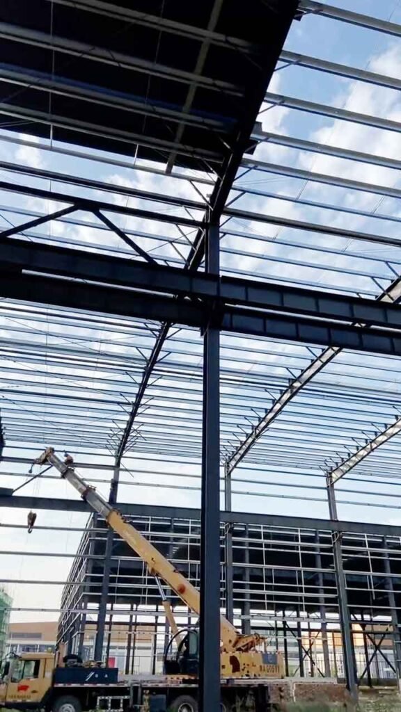 Grúa telescópica instalando vigas de acero en una bodega de marco metálico de gran altura y luces libres amplias.