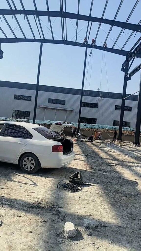 Construction site of a heavy industrial warehouse showing the large-scale steel skeleton and high-bay portal frames with a vehicle for scale.