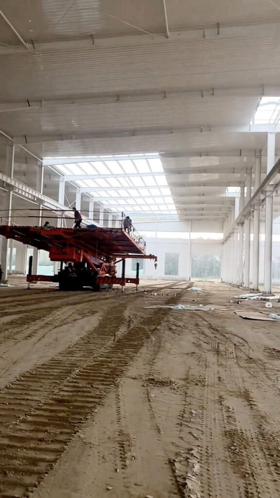 Ongoing construction site of a large warehouse structure with a mobile scissor lift and ground preparation for a high-strength concrete radier