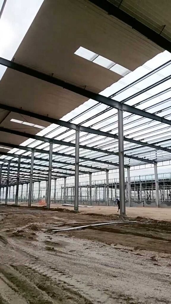 Ground-view of a tropical warehouse structure with reflective roofing sheets and a portal frame skeleton under construction.