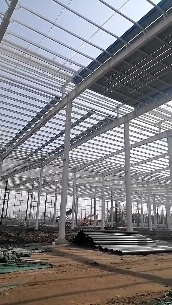 Roof panel installation over a rigid steel frame with raw materials staged below, representing the comprehensive material packages delivered by an industrial steel building supplier.