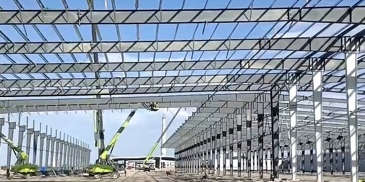 Dual cranes operated by an industrial warehouse contractor lifting massive Q355 structural steel rafters into place for a multi-span logistics facility.