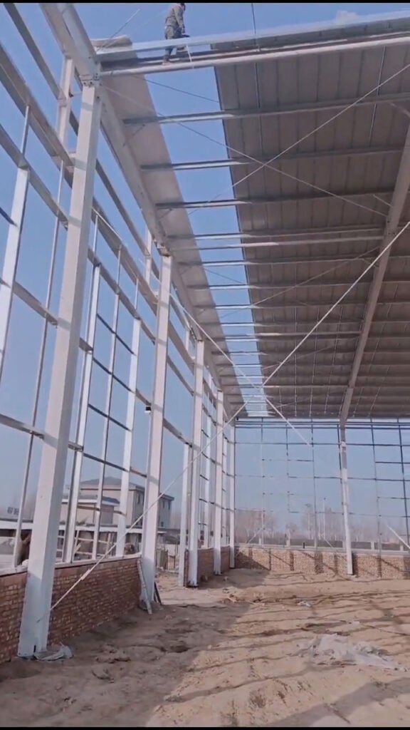 Construction workers installing galvanized roof purlins on a steel structure provided by a certified steel building kit supplier, integrating with a traditional brick half-wall.