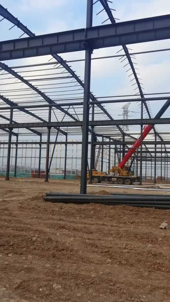Heavy-duty mobile crane installing primary steel columns and roof beams on an industrial steel warehouse site, supplied by a China steel warehouse manufacturer.