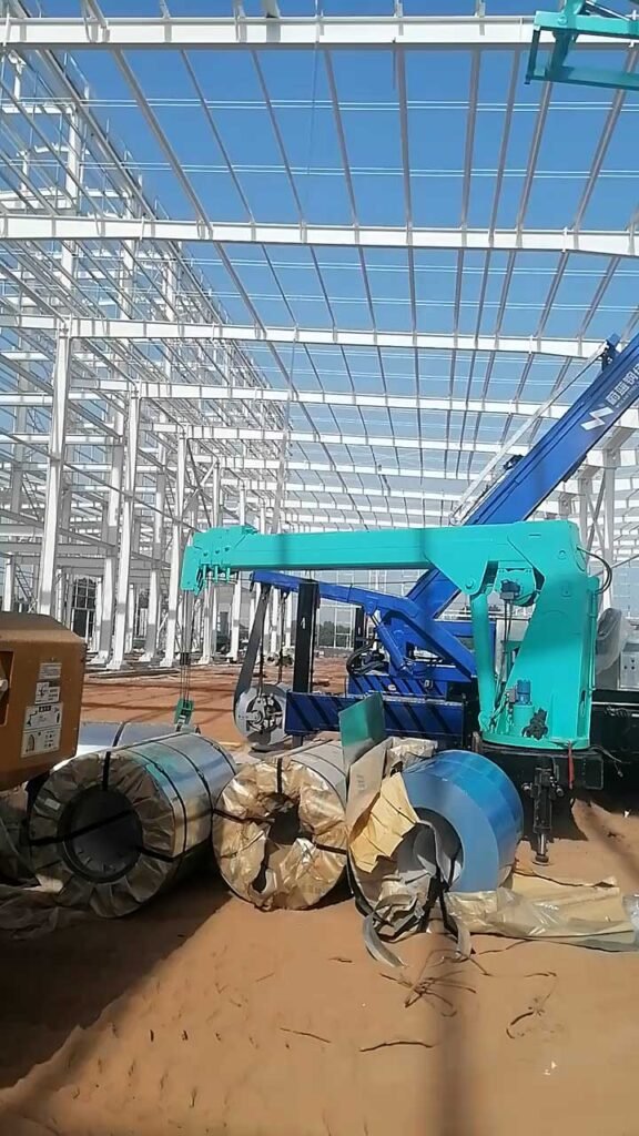 crane lifting steel coils next to an erected structural frame, demonstrating the heavy machinery requirements often factored into a comprehensive steel warehouse quotation