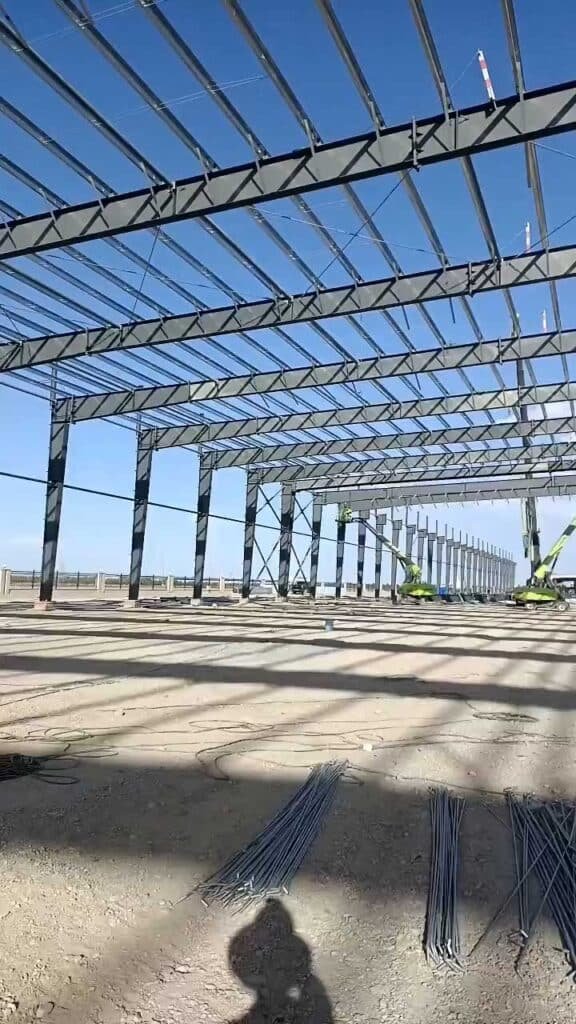 A professional warehouse building contractor managing the installation of galvanized roof purlins and cross-bracing on a large-span portal frame structure under a clear blue sky.