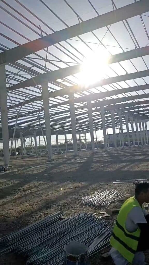 Site worker organizing anchor bolts and rebar in the foreground, with the massive structural steel framework erected by a certified warehouse construction company in the background.