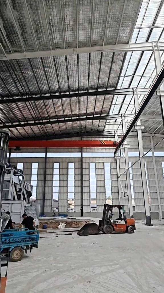 Estructura de acero de almacén industrial con puente grúa rojo ensamblado y montacargas operando en la base